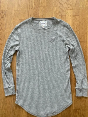 Grå ribbad långärmad tröja SikSilk M - Säljer en grå ribbad långärmad tröja från SikSilk. Tröjan har rund halsringning, diskret broderad logga på bröstet och mjukt material som känns skönt mot huden. Perfekt för en avslappnad och stilren look.