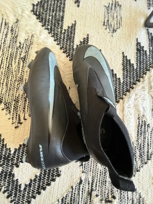 Svarta Nike fotbollsskor Air Zoom - Säljer ett par svarta Nike fotbollsskor med silverdetaljer och Air Zoom-tryck på hälen. Skorna har en strömlinjeformad design utan snörning och är tillverkade i syntetmaterial för bästa grepp och kontroll på planen. Perfekta för dig som vill ha stilrena och funktionella skor. Jag köpte dem för ett halvår sedan men märkte att dem inte passade mig helt. Sen dess har dem varit i en låda och därför dem inte ser helt nya ut. Det ingår speciella sulor med dessutom. Hör ut om du vill förhandla om prise