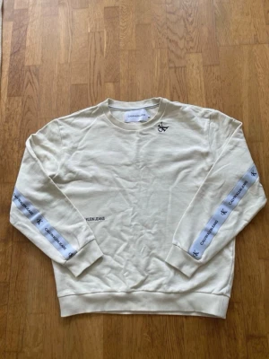 Vit sweatshirt från Calvin Klein Jeans - Vit sweatshirt med rund hals och långa ärmar från Calvin Klein Jeans. Tröjan har logoband med CK-tryck längs ärmarna och över ryggen samt liten logga på bröstet. Mjuk bomullskänsla och ribbade muddar.