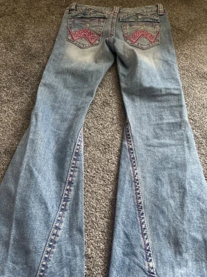 Ljusblå bootcut jeans med brodyr - Säljer ett par ljusblå bootcut jeans med snygg brodyr i rosa och rött på bakfickorna samt längs bensömmarna. Jeansen har låg midja och klassisk femficksdesign. Tillverkade i denim med en cool tvättad look och unika detaljer.
