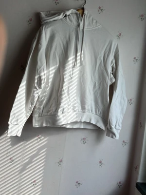 Vit hoodie med huva och dragsko - Säljer en enkel och stilren vit hoodie med huva och dragsko. Hoodien har långa ärmar, ribbade muddar och en avslappnad passform. Perfekt för dig som gillar en clean och basic look. Materialet känns mjukt och bekvämt, troligen bomull eller bomullsblandning.