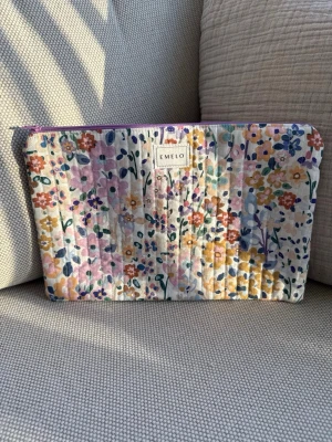 Blommig quiltad necessär från Emelo - Säljer en quiltad necessär med färgglatt blommönster i rosa, gult, blått och grönt. Väskan har dragkedja upptill och är perfekt för småsaker eller smink. Märket Emelo syns framtill. Materialet känns mjukt och tygigt. Mått ca 29x20cm