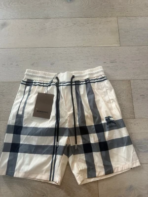 Rutiga shorts från Burberry med snörning - Snygga vita shorts från Burberry med klassiskt rutigt mönster i svart och grått. Shortsen har elastisk midja med snörning och Burberry-logga på ena benet. Perfekta för sommaren och ger en lyxig känsla till din outfit.