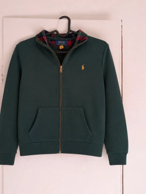 Ralph Lauren zip tröja - Säljer en riktigt snygg vintage zip-up från Polo Ralph Lauren i mörkgrönt med klassisk gul logga och unika rutiga detaljer på insidan av kragen. En tidlös modell som är svår att hitta idag och som passar perfekt för dig som gillar vintage och preppy-stilen. Tröjan är i storlek M (10–12) och har en bekväm passform med dragkedja framtill samt två praktiska fickor. Den är i mycket bra skick och har inga fläckar eller skador😁