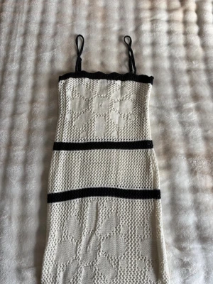 Vit lång stickad klänning med svarta detaljer - Säljer en vit lång stickad klänning med svarta axelband och svarta ränder. Klänningen har ett blommigt mönster i stickningen och är ärmlös med smala axelband. Perfekt för sommarens alla tillfällen.