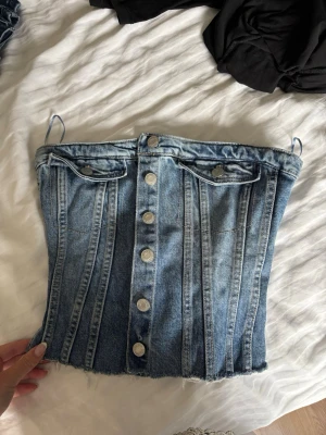 Jeans korsettopp med knappar - Säljer en snygg jeans korsettopp med knappar framtill och dekorativa fickor. Toppen har smala axelband och rå kant nedtill för en trendig look. Perfekt att styla med höga byxor eller kjol.