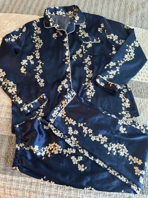 Mörkblå blommig satinpyjamas XL  - Snygg tvådelad pyjamas i mörkblå satin med vita blommor och vita kantdetaljer. Skjortan har knappar framtill, bröstficka och långa ärmar. Byxorna har resår i midjan för extra komfort. Perfekt för mysiga hemmakvällar.