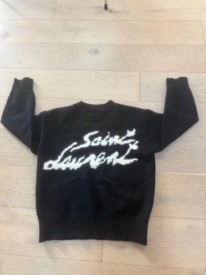 Svart Saint Laurent stickad tröja - Svart stickad tröja från Saint Laurent med vit logotyp i grafisk stil på bröstet. Tröjan har rund halsringning, långa ärmar och ribbstickade muddar vid ärmslut och nederkant. Perfekt för dig som gillar streetwear och exklusiva märken.