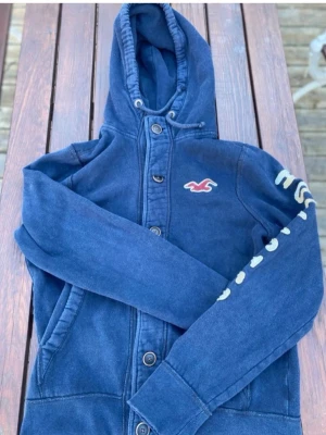 Mörkblå cardigan från hollsitet - Mörkblå hoodie från Hollister med dragkedja och knappar framtill. Har huva med dragsko, fickor på sidorna och Hollister-logga på bröstet samt tryck på ena ärmen. Perfekt för en avslappnad och cool stil. Storlek på lappen står l men den är som m⚠️