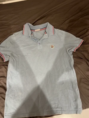 Ljusblå Moncler pikétröja - Snygg ljusblå pikétröja från Moncler med klassisk krage och korta ärmar. Tröjan har rödvita detaljer på krage och ärmslut samt Moncler-logga på bröstet. Perfekt till en avslappnad men stilren look.