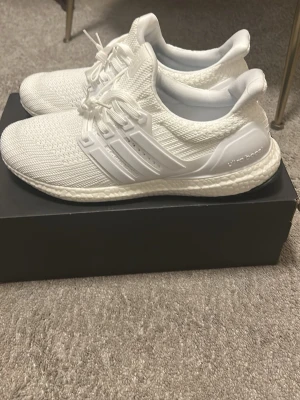 Helvita Adidas Ultraboost sneakers - Storlek 45. Säljer ett par helvita Adidas Ultraboost sneakers med stickad ovandel och klassiska tre ränder på sidan. Skorna har en bekväm och responsiv Boost-sula samt vita snören och detaljer. Perfekta för dig som gillar stilrena och sportiga sneakers.