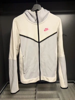 Vit och ljusgrå Nike hoodie - Snygg hoodie från Nike i vitt och ljusgrått med dragkedja framtill och huva. Har svarta detaljer och en rosa Nike-logga på bröstet. Tillverkad i mjukt material och har fickor på sidorna. Perfekt för en sportig och avslappnad stil. Använd 1,2 gånger