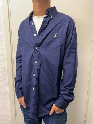 Ralph Lauren skjorta - Snygg mörkblå skjorta med klassisk krage och vita knappar. Skjortan har lång ärm och en gul broderad detalj på bröstet. Perfekt till jeans för en stilren look.