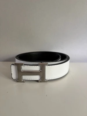 Vitt skärp med silverfärgat H-spänne - Snyggt vitt skärp med ett stort, silverfärgat H-format spänne. Skärpet har en stilren design och är gjort i slätt skinn. Insidan är svart vilket ger en cool kontrast. Perfekt accessoar för att lyfta din outfit.
