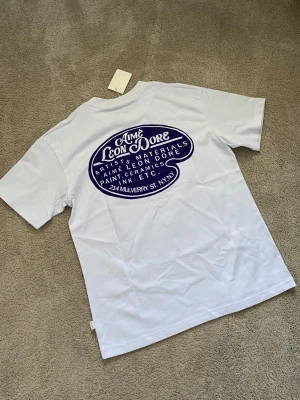 Vit Aimé Leon Dore t-shirt med tryck - Säljer en vit t-shirt från Aimé Leon Dore med blått tryck både fram och bak. T-shirten har klassisk passform, rund hals och korta ärmar. Tillverkad i mjuk bomull och har en stilren, modern vibe.