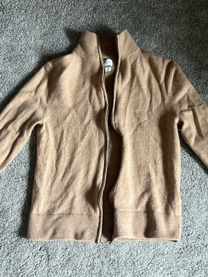 Beige merinoullkofta från These Glory Days - Snygg beige kofta i merinoull från These Glory Days. Dragkedja framtill, ribbade muddar och hög krage ger en clean look. Perfekt för lager på lager och riktigt skön tack vare det mjuka materialet i Superior Merino. Står L. Men är väldigt liten i storleken. Uppskattningsvis XS