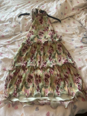 Blommig volangklänning med halterneck - Säljer en söt kortklänning med blommigt mönster i grönt, rosa och beige. Klänningen har halterneck, flera volanger och är ärmlös. Perfekt för sommarens alla tillfällen!