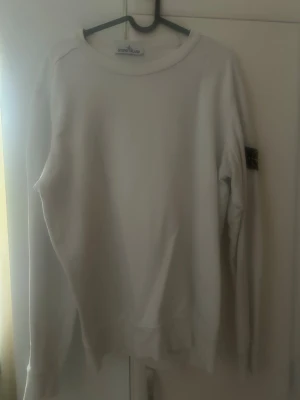 Vit sweatshirt från Stone Island - Vit sweatshirt från Stone Island med klassisk rund hals och den ikoniska svarta patchen, Finns en liten fläck på ena armen kan försvinna i tvätten har ej testa, tröjan har en clg kod som går att scanna för att få bekräftat att den är äkta 
