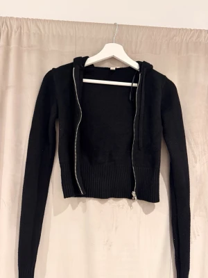 Svart croppad hoodie med dragkedja - Säljer en svart croppad hoodie med dragkedja framtill och ribbad kant nedtill. Hoodien har långa ärmar och en klassisk huva. Materialet är stickat och ger en mjuk känsla. Perfekt för en chill och trendig look.