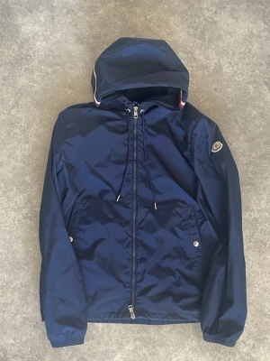 Moncler vindjacka  - Snygg mörkblå vindjacka från Moncler med huva och dragsko. Jackan har två fickor med tryckknappar, hel dragkedja framtill och Moncler-logga på ärmen. Materialet är glansigt och lätt, perfekt för blåsiga dagar.