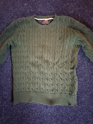 Grön kabelstickad tröja Polo Ralph Lauren - Mysig grön kabelstickad tröja från Polo Ralph Lauren med rund halsringning och ribbade muddar. Tröjan har det klassiska lilla broderade logotypen i lila på bröstet. Perfekt för kyliga dagar och enkel att matcha med jeans.