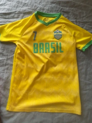 Gul Brasil fotbollströja H&M - Snygg gul fotbollströja från H&M med gröna detaljer, Brasil-tryck och nummer 7 på bröstet. Tröjan har rund halsringning och är tillverkad i ett lätt och svalt material, perfekt för träning eller match.