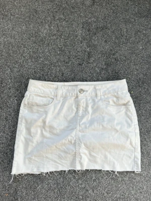 Vit jeanskjol med rå kant - Snygg vit jeanskjol i minilängd med fem fickor och rå, fransig kant nedtill. Kjolen har knapp- och dragkedjestängning framtill samt bälteshällor. Perfekt till sommaren och enkel att matcha med olika toppar.