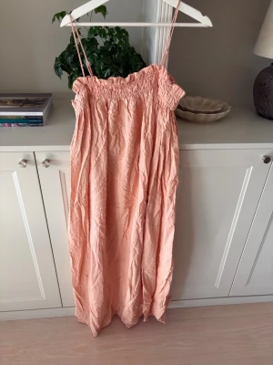 Ljusrosa långklänning med smock - Säljer en ljusrosa långklänning med smala axelband och smock upptill. Klänningen har en avslappnad passform och är tillverkad i ett lätt, skrynkligt tyg som passar perfekt till varma dagar. Enkel och stilren design som ger en somrig känsla.