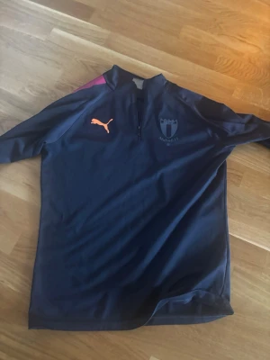 Mörkblå Malmö FF träningströja Puma - Snygg mörkblå träningströja från Puma med Malmö FF-logga på bröstet och orange Puma-logga. Tröjan har halv dragkedja framtill och lila detalj på axeln. Perfekt för fotbollsträning eller gymmet.