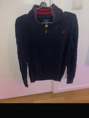 Ralph Lauren quater zip - Mörkblå kabelstickad tröja från Polo Ralph Lauren med halv dragkedja och hög krage. Röd broderad logga på bröstet och röd randig detalj på insidan av kragen. Perfekt för en stilren och klassisk look.