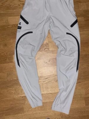 Gråa mjukisbyxor Under Armour - Snygga gråa mjukisbyxor från Under Armour med svarta detaljer och dragkedjefickor. Byxorna har resår i bensluten och en sportig look med tryck på ena benet. Perfekta för chill eller träning.
