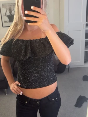 Glittrig svart offshoulder topp - Säljer en glittrig svart offshoulder topp med volang runt axlarna. Toppen är croppad och har en tight passform som framhäver figuren. Perfekt för dig som vill sticka ut lite extra med en snygg och trendig look. Använd 1 gång!