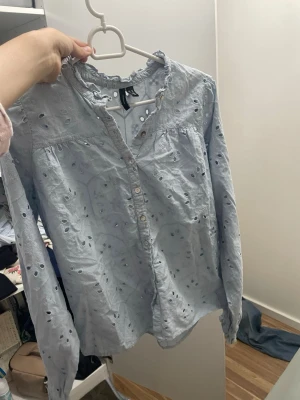 Ljusblå broderad blus med volang - Söt ljusblå blus med broderade hål och volangkant vid halsen. Blusen har knappar framtill och långa ärmar med resår vid ärmslut. Tillverkad i ett tunt, luftigt bomullstyg som ger en romantisk och trendig look.