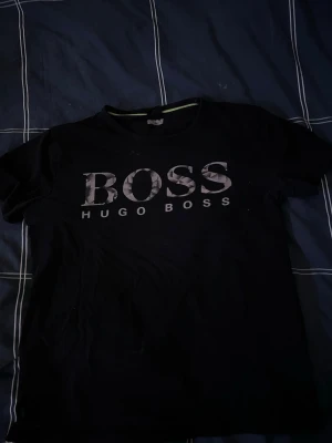Svart t-shirt från Hugo Boss - Snygg svart t-shirt från Hugo Boss med stort BOSS-tryck i grått och vitt på bröstet. Klassisk rund hals och korta ärmar. Perfekt för dig som gillar stilrena och trendiga plagg med tydlig logga.