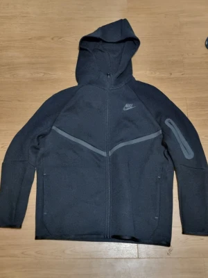 Svart Nike hoodie med dragkedja - Snygg svart hoodie från Nike med hel dragkedja framtill och huva. Har gråa detaljer över bröstet och en ficka med dragkedja på ena ärmen. Säljer den för den är för stor. Aldrig andvänd. Den är mörkare irl. 170cm