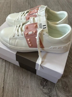 Valentinos - Snygga vita sneakers med breda rosa band över vristen dekorerade med matchande nitar. Skorna har vita snören, platt sula och detaljerade sömmar. Perfekta för dig som vill sticka ut med en cool och trendig look. Strl 38 men passar 37 skulle jag säga💕