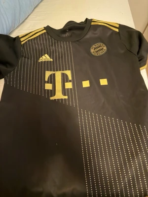 Svart Bayern München matchtröja - Snygg svart matchtröja från Bayern München med guldiga detaljer och Adidas-logga. Tröjan har korta ärmar, guldfärgade ränder på axlarna och ett stort T-mobil-tryck på bröstet. Perfekt för fotbollsträning eller att bära till vardags.