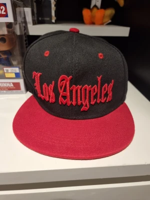 Svart och röd Los Angeles keps - INSTABOX!!! Snygg snapback-keps i svart med röd skärm och broderad text 'Los Angeles' i rött framtill. Kepsen har platt skärm, ventilationshål och justerbar plastrem bak. Perfekt för dig som gillar streetwear och vill sticka ut.
