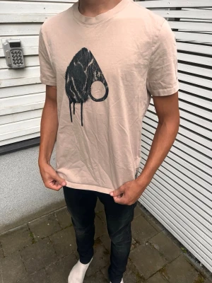 Moose knuckles t-shirt - Säljer en beige t-shirt med ett stort svart grafiskt tryck på bröstet. T-shirten har rund halsringning och korta ärmar. Materialet känns mjukt och skönt, perfekt för en avslappnad stil.