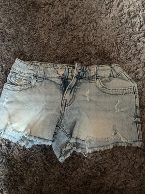 Ljusblå slitna jeansshorts - Säljer ett par ljusblå jeansshorts med slitna detaljer och råa kanter. Shortsen har fem fickor, bälteshällor och snygga broderade bakfickor. Perfekta för varma dagar och en avslappnad stil. Det är storlek 158 och köpta från Gina tricot  