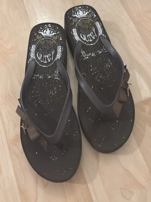 Bruna sandaler från Juicy Couture - Snygga bruna sandaler från Juicy Couture med vita stänk och dekorativt tryck på sulan. Remmarna har en brun rosett och ett litet hänge med Juicy Lovers-logga. Perfekta för en trendig och avslappnad look.