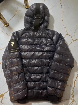 Mörkgrå dunjacka med huva - Snygg mörkgrå dunjacka från Peak Performance med stor huva och breda, vadderade paneler. Jackan har dragkedja framtill, ribbade muddar och en cool detalj med spänne på huvan. Märkeslogga på ärmen och två sidofickor med dragkedja.