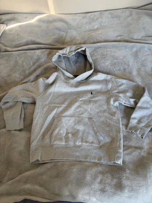 Grå hoodie från Polo Ralph Lauren - Snygg grå hoodie från Polo Ralph Lauren med klassisk huva och känguruficka framtill. Liten mörkblå logga broderad på bröstet. Tillverkad i mjukt bomullsmaterial med ribbade muddar vid ärmslut och nederkant. Perfekt för en avslappnad och stilren look.