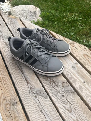 Grå adidas skor   med svarta ränder - Snygga grå sneakers från Adidas med klassiska svarta ränder på sidan. Skorna har lågt skaft, grå snörning och vit sula med svart rand. Tillverkade i syntetmaterial och textil för en clean och sportig look.
