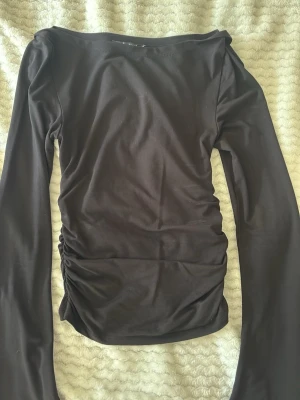 Svart långärmad topp Gina Tricot - Säljer en svart långärmad topp med boat Neck från Gina Tricot i stretchigt material. Toppen har en tight passform och snygg rynkning på båda sidorna, som ger en cool detalj. Perfekt att styla med jeans eller kjol för en trendig look.