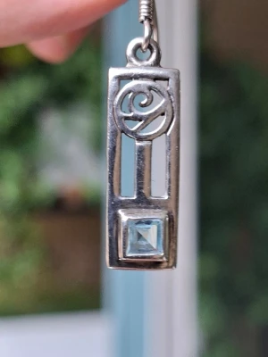 Silver, Sterling 925, örhängen från Kit Heath Jewellery, England - Retroörhängen i Sterlingsilver med slipad topas i himmelsblått. Designen är inspirerad av Art nouveauarkitekten Charles Rennie Mackintosh's ornamentik från 1900-talets början.    Plattans längd är 22 mm och bredden 18 mm. Örhängets totala längd är ca 40 mm. Örhängena är köpta på Kit Heath Jewellery i England kring 1990-talet och har aldrig använts. Mycket gott skick! Originalasken medföljer.                             