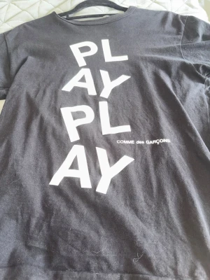 Svart PLAY t-shirt Comme des Garçons - Cool svart t-shirt från Comme des Garçons med stort vitt PLAY-tryck framtill. Klassisk rund hals och korta ärmar. Perfekt statement-plagg för dig som gillar streetwear och vill sticka ut.