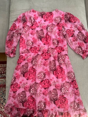 Rosa blommig långklänning - Säljer en rosa långklänning med blommigt mönster i olika nyanser av rosa och lila. Klänningen har v-ringning, volangdetaljer och trekvartsärm. Perfekt för dig som vill sticka ut med färg och mönster. Materialet känns lätt och luftigt.