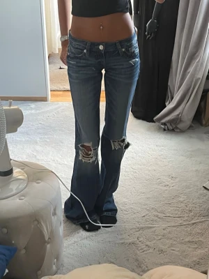 Blå slitna bootcut jeans - Säljer ett par blå bootcut jeans med slitningar och hål vid knäna. Jeansen har låg midja och klassisk femficksmodell. Materialet är denim med en tvättad look och råa detaljer för en trendig vibe. Använda 1 gång! Storlek xxs/tall!!