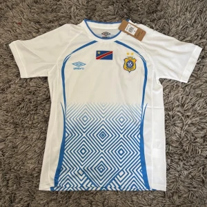 Vit fotbollströja från Umbro ECOFF - Snygg vit fotbollströja från Umbro med blå detaljer och grafiskt mönster nedtill. Tryck av flagga och ECOFF-emblem på bröstet. Perfekt för match eller träning. Tillverkad i lätt och ventilerande material.